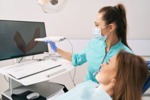 Female stomatologist holding intraoral dental scanner and pointing at computer display while discussing dental treatment with patient